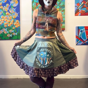 May include: A person wearing a multi-colored crop top, a green skirt with a "Protect Our Planet" patch, and a burgundy hat. The skirt has a ruffled hem with a patterned border. The person is wearing a mask and jewelry.
