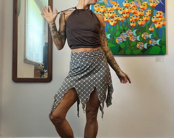 May include: A woman wearing a brown halter top and a black and white patterned skirt with a unique, asymmetrical design. The skirt has multiple slits and a fringed hem. She is wearing black shoes. A colorful painting of fish is on the wall behind her.