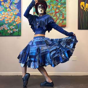 May include: A blue long-sleeve cropped top with ruffled shoulders and a matching blue patchwork skirt featuring multiple layers of fabric strips in various shades of blue. The outfit is paired with a dark hat and a white and black patterned face mask.