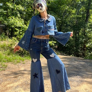 May include: A denim outfit featuring a cropped jacket and flared jeans. The jacket has bell sleeves and embroidered details. The jeans have star and heart patches. The outfit is a light blue wash, with a vintage aesthetic.