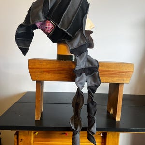 May include: A handmade black and gray jester hat with a long, spiraling scarf. The hat is constructed from various fabric pieces and features a purple and pink patterned lining. The hat is displayed on a mannequin head.