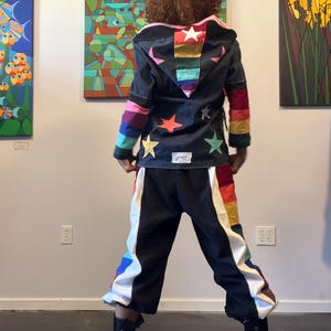 May include: A black hooded jacket and matching trousers featuring colourful star appliqués and rainbow-striped panels. The jacket has a hood with a rainbow centre stripe and a white rectangular label at the hem. The trousers have wide white side panels.