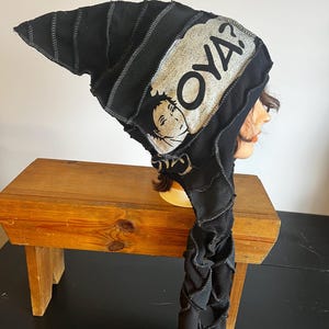 Pode incluir: Chapéu preto pontudo com a palavra "OYA?" e um gráfico de um rosto. O chapéu é feito de tecido costurado e tem uma longa cauda decorativa. O chapéu é exibido em um banco de madeira.