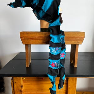 Peut inclure: Un chapeau fantaisiste fait à la main avec une longue queue flottante. Le chapeau présente des panneaux de tissu noirs et bleu turquoise alternés. La queue est ornée de motifs floraux brodés. Le chapeau est présenté sur une tête de mannequin.