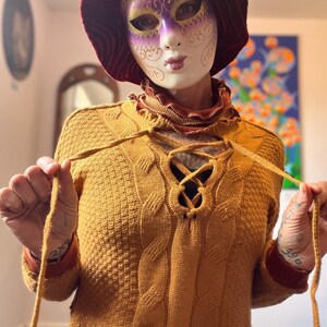 Puede incluir: Jersey de punto amarillo mostaza con escote con cordones y detalles de punto trenzado. El jersey tiene mangas largas con puños burdeos y un dobladillo burdeos. Se lleva una máscara decorativa con detalles morados y dorados.