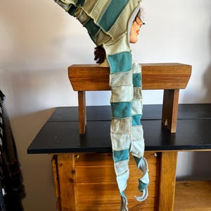May include: A patchwork hood and scarf set in shades of teal, beige, and cream. The hood is elongated and pointed, with a matching scarf that drapes down. The fabric appears to be a soft, textured material, possibly fleece or a similar textile.