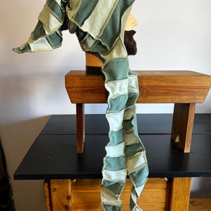 May include: A green and cream-colored patchwork hat with a long, flowing tail. The hat is made of fabric strips sewn together, creating a striped pattern. The hat is displayed on a mannequin head.
