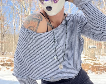 May include: A person wearing a gray and black striped sweater with a white mask with purple and blue accents. The mask has a star and floral design. The person is wearing a black necklace with a silver pendant. The person is standing in a snowy forest.