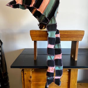 May include: A patchwork hat with a long, trailing scarf. The hat and scarf are made of fabric strips in black, pink, and green, sewn together with visible stitching. The hat is displayed on a mannequin head.