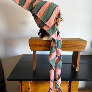 Peut inclure: Un chapeau artisanal fantaisiste avec une longue queue en spirale. Le chapeau présente des rayures alternées de tissu rose poudré et vert, cousues ensemble avec des coutures visibles. Le chapeau est présenté sur une tête de mannequin.