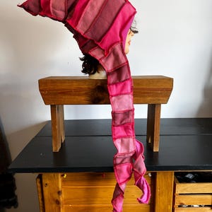 May include: A patchwork hood in shades of pink and red, with a long, spiraling tail. The hood is made of rectangular fabric pieces sewn together. The headwear is displayed on a mannequin head.