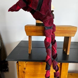 May include: A handmade, whimsical hat in shades of burgundy and black, featuring a long, twisted design. The hat is constructed from textured fabric and has a pointed tip. It is displayed on a mannequin head, showcasing its unique shape and style.