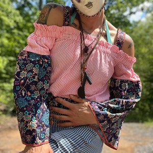 May include: A person wearing a pink and white striped off-the-shoulder top with floral print sleeves. The top has a ruffled neckline and the sleeves are bell-shaped. The person is also wearing a blue and white striped skirt and a white mask with purple and gold accents.