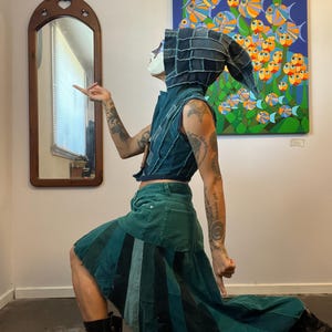 Puede incluir: Un atuendo de mezclilla patchwork que consiste en un chaleco y una falda larga. La falda es una mezcla de paneles de mezclilla verdes y azules. La persona lleva una capucha de mezclilla y botas negras. Una pintura colorida de peces cuelga en la pared.