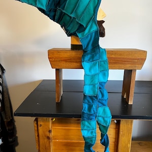 Pode incluir: Um capuz e cachecol de patchwork em tons de azul e turquesa. O capuz é pontudo e o cachecol é longo e retangular, com uma série de quadrados costurados. O artigo é exibido em uma cabeça de manequim.