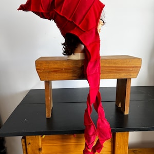 May include: A vibrant red, pointed hat with a long, flowing tail. The hat is constructed from multiple fabric panels, creating a textured, layered appearance. The hat is displayed on a mannequin head, showcasing its shape and design. The hat is likely a costume piece.