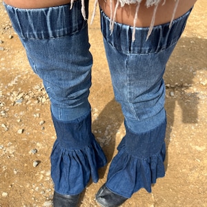 Può includere: Un paio di scaldagambe in denim con un fondo a balze. Gli scaldagambe sono realizzati in tessuto denim blu scuro e hanno un elastico elastico in alto.