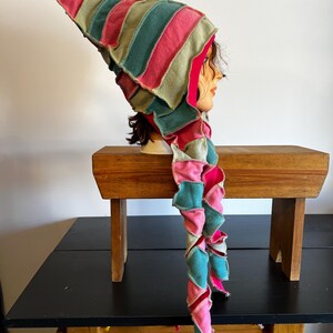 May include: A colorful, patchwork hat with a long, spiraling tail. The hat features alternating stripes of pink, green, and teal fabric. The tail is made of the same patchwork material, creating a whimsical and unique accessory.