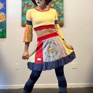 May include: A colorful outfit featuring a yellow crop top with red trim and color-blocked sleeves. The skirt is patchwork with the text "THE WORLD NEEDS ALL KINDS OF MINDS". The outfit is paired with dark boots and leggings.