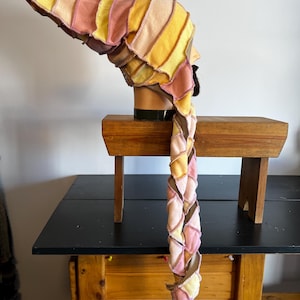May include: A colorful, patchwork hat with a long, braided tail. The hat features a series of fabric panels in shades of yellow, pink, and brown. The hat is displayed on a mannequin head, showcasing its unique design and construction.