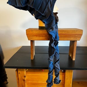 Op de afbeelding: Een donkerblauwe en grijze patchwork kap met lange, gedraaide sjaalachtige uiteinden. De kap wordt getoond op een mannequinhoofd, waarbij het unieke, gelaagde ontwerp wordt getoond. De stof lijkt een zacht, getextureerd materiaal te zijn.