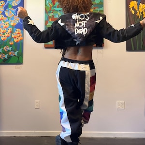 May include: A black cropped denim jacket with white star patches and a hood featuring the text EMO'S NOT DEAD in white, paired with black and white color-blocked joggers featuring vertical rainbow-colored side panels and black boots.