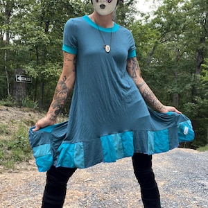 Può includere: Una donna indossa un vestito blu e turchese con un orlo a patchwork. Indossa una maschera bianca con un motivo a farfalla e una collana d'argento. Il vestito ha maniche corte e una scollatura rotonda. La donna è in piedi su un sentiero di ghiaia in un bosco.