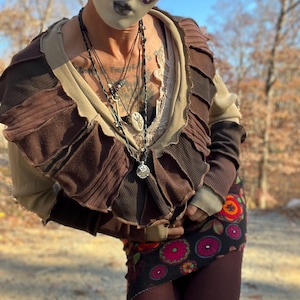 Op de afbeelding: Een persoon draagt een bruin en beige gelaagd topje met een gerimpelde halslijn. Ze draagt een zwarte en rode rok met een bloemenpatroon en bordeauxrode leggings. De persoon draagt ook een wit masker met paarse accenten en meerdere kettingen.