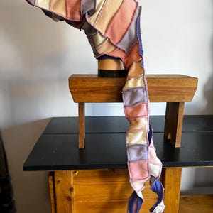 Pode incluir: Um cachecol colorido em patchwork em tons pastel de rosa, roxo, pêssego e creme. O cachecol é feito de pedaços retangulares de tecido costurados. O cachecol tem uma cauda longa e fluida com detalhes azuis e brancos. O cachecol é exibido em uma cabeça de manequim.