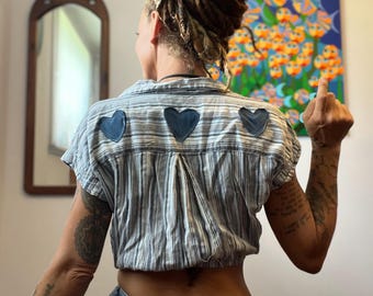May include: A cropped, short-sleeved shirt with blue and white stripes and three dark blue heart-shaped patches on the back. The shirt is paired with light blue denim jeans. The image is taken indoors.