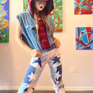 May include: A denim vest and star-patched jeans outfit. The vest is sleeveless and layered over a burgundy top. The jeans feature large white and blue stars. A hooded head covering with a red and gray pattern is worn.