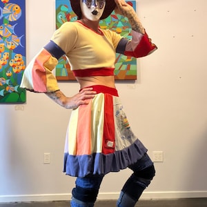 May include: A yellow crop top with red trim and colorful bell sleeves is paired with a patchwork skirt and blue jeans. The skirt features a variety of colors and a ruffle hem. A dark hat and mask complete the look.