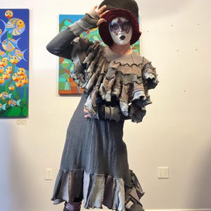 May include: A theatrical costume. The outfit includes a grey dress with layered ruffles and patchwork details, a matching top, striped leggings, and a large black hat with a burgundy brim. A decorative mask completes the look.