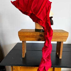 May include: A vibrant red medieval-style hat with a long, flowing tail. The hat is made of a soft fabric and features vertical pleats on the main portion. It is displayed on a mannequin head with a beard, sitting on a wooden bench.