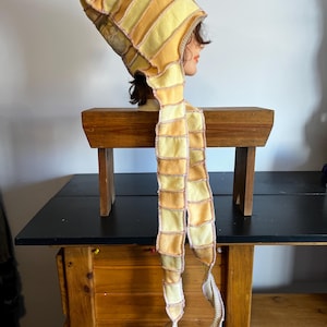 May include: A unique, handmade hat with a long scarf-like extension. The hat and scarf are made of yellow and orange fabric, stitched together in a patchwork design. The hat is displayed on a mannequin head.