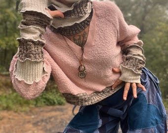 May include: A person wearing a pink sweater with a brown and white knit cowl neck, a gold and purple mask, and blue patchwork pants. The person is wearing brown and white knit fingerless gloves.