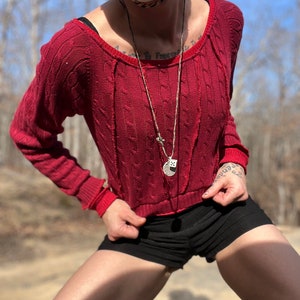 May include: A person wearing a red knitted jumper with a white mask and a silver necklace with a dice charm. The person is wearing black shorts and has a tattoo on their arm.