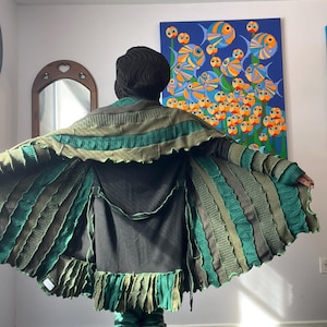 May include: A person wearing a handmade green and brown knitted cape with a black underlayer. The cape features a unique design with layered textures and fringe detailing. The person is also wearing matching leg warmers.