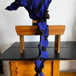 Könnte beinhalten: Ein handgefertigter Hut mit langem, fließendem Schwanz. Er hat abwechselnd schwarze und blaue Fleece-Streifen. Der Hut wird auf einem Schaufensterkopf präsentiert. Das Design des Hutes ist skurril und einzigartig.
