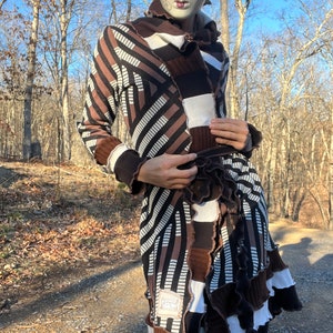 May include: A long brown and white striped cardigan with a ruffled hem and a belt. The cardigan is made of a soft, knitted fabric and has a relaxed fit. The model is wearing a masquerade mask.