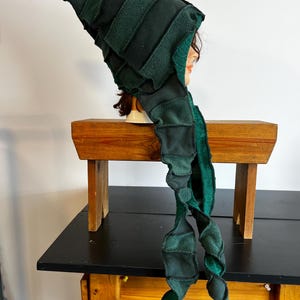 Pode incluir: Um chapéu de lã verde escuro com uma longa e fluida echarpe. O chapéu tem um topo pontudo e é feito de várias tonalidades de lã verde, criando um efeito em camadas. A echarpe é uma continuação do design do chapéu, com uma aparência ondulada e texturizada.
