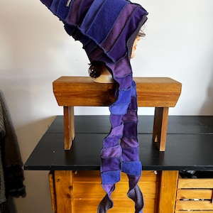 Peut inclure: Ensemble fait main composé d'un chapeau et d'une écharpe en patchwork dans les tons de violet et de bleu. Le chapeau a un sommet pointu et une longue écharpe flottante attachée. Le tissu semble être une matière douce et texturée, peut-être de la polaire ou de l'éponge. L'ensemble est présenté sur une tête de mannequin.