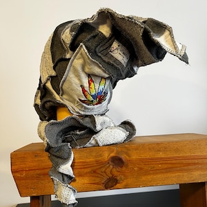 May include: A patchwork hat with a colorful cannabis leaf design. The hat is made from various shades of gray and white fabric, with a ruffled edge. The hat is displayed on a wooden bench.