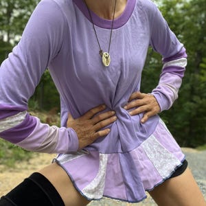 Könnte beinhalten: Ein langärmeliges, violettes Shirt mit einem Rüschen-Saum. Das Shirt hat ein weiß-violettes Streifenmuster an den Ärmeln und einen weißen und violetten Musterbesatz am Saum. Das Shirt wird von einer Person mit einer weißen Maske und einer Halskette mit einem Anhänger getragen.