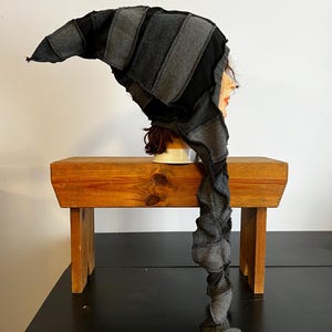 May include: A handmade, patchwork hat with a long, spiraling tail. The hat is constructed from various shades of gray fabric, including denim and other textiles. The hat is displayed on a mannequin head.
