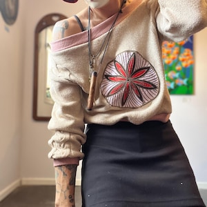 以下が含まれることがあります： 前面に赤と白の花柄模様が入ったベージュ色のセーター。セーターは、ネックラインと袖口にピンク色のトリムが付いています。黒いスカートとマスク付きの帽子も確認できます。