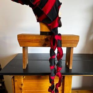 Pode incluir: Um chapéu de patchwork feito à mão em vermelho e preto. O chapéu tem uma longa cauda torcida e é exibido em uma cabeça de manequim. O chapéu é feito de tecido e tem um topo pontudo. O chapéu está em uma mesa de madeira.