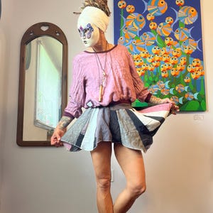 May include: A person wearing a pink long-sleeved top with a dark burgundy trim and a patchwork skirt. The person is wearing a white mask with a decorative design and a long necklace. A painting of colourful fish hangs on the wall.