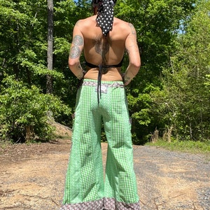 May include: Green and white checkered wide-leg pants with a brown and white patterned trim. The pants have a tie-waist and are worn with a black top and a black and white polka dot headscarf.