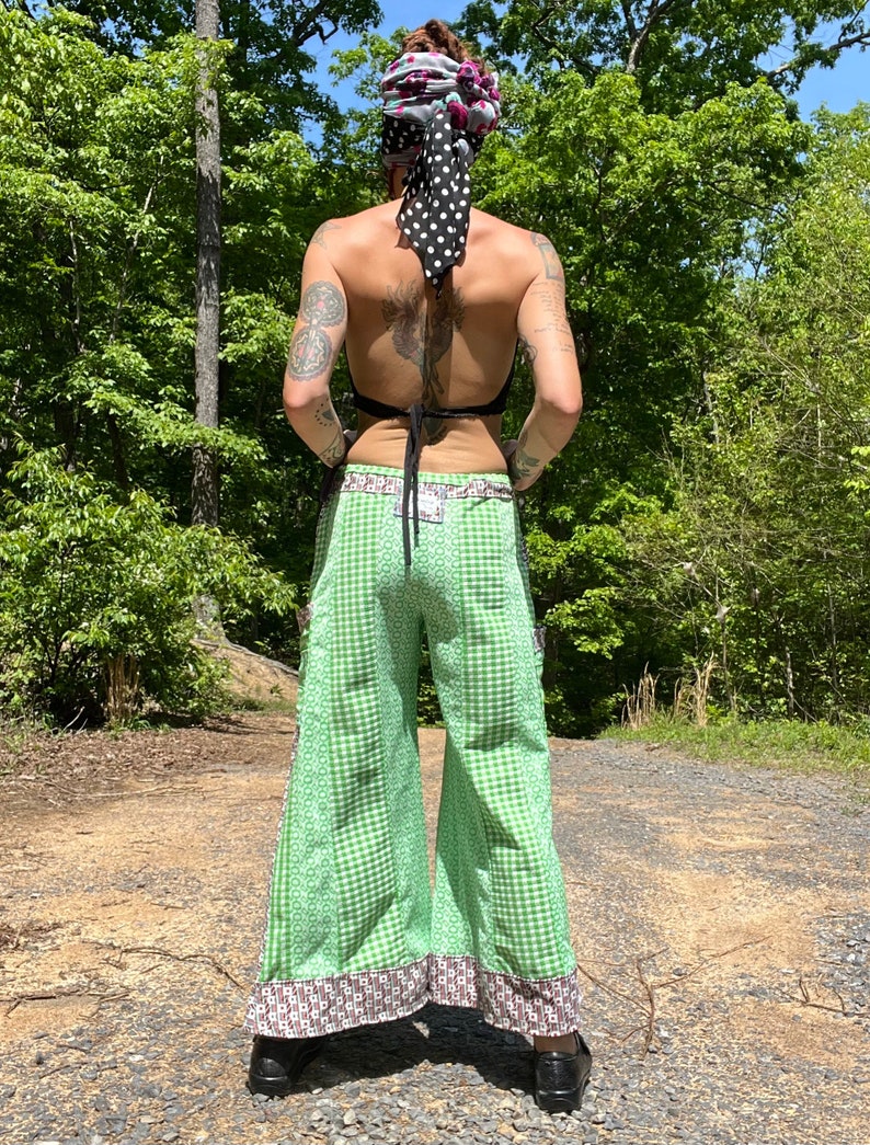 May include: Green and white checkered wide-leg pants with a brown and white patterned trim. The pants have a tie-waist and are worn with a black top and a black and white polka dot headscarf.